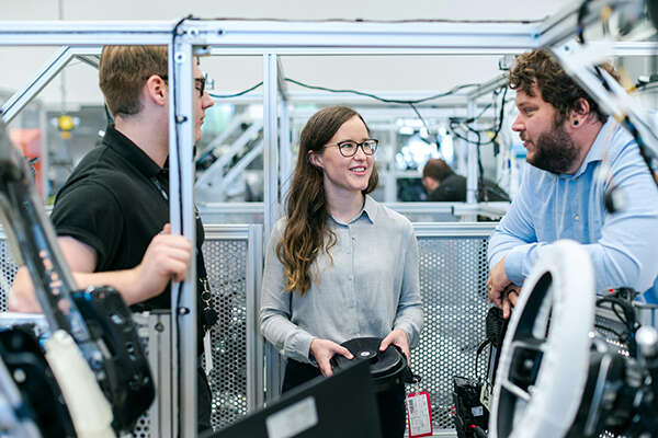 A smiling female engineering apprentice flanked by two males in a carmaking factory